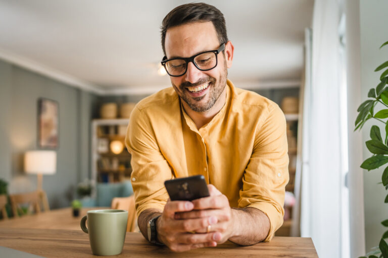 Ein Mann lehnt in entspannter Pose über einen Tisch gebeugt und blickt mit glücklichem Gesichtsausdruck auf sein Smartphone